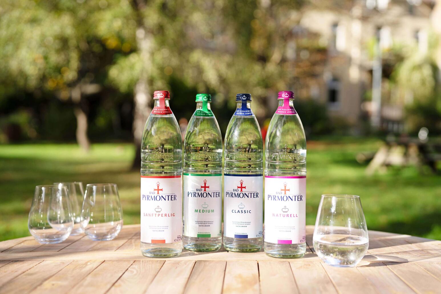 Vier Bad Pyrmonter Mineralwasserflaschen in verschiedenen Sorten stehen auf einem Holztisch im Freien, daneben leere Gläser.