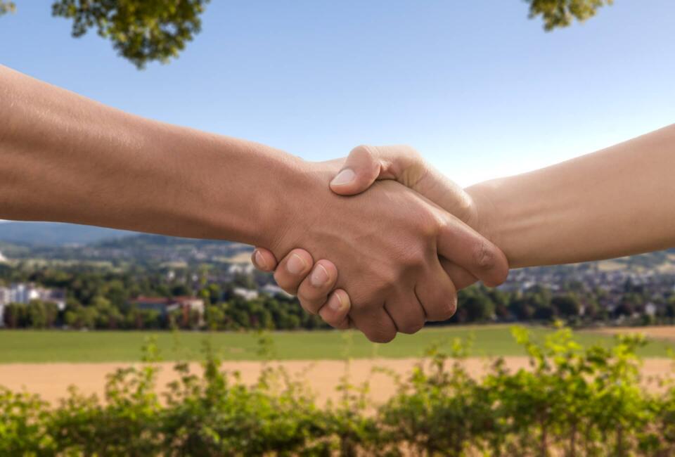 Zwei Hände geben sich vor Landschaft und blauem Himmel einen Handschlag.