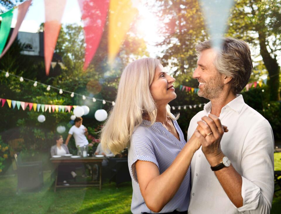 Älteres Paar tanzt glücklich im Garten bei einer Feier mit Lichterketten und Wimpeln.