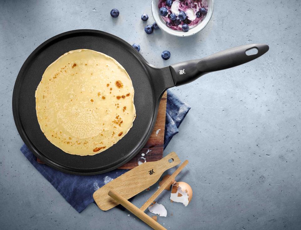 Pfannkuchen in einer schwarzen Pfanne mit Kochutensilien und Blaubeeren im Hintergrund.
