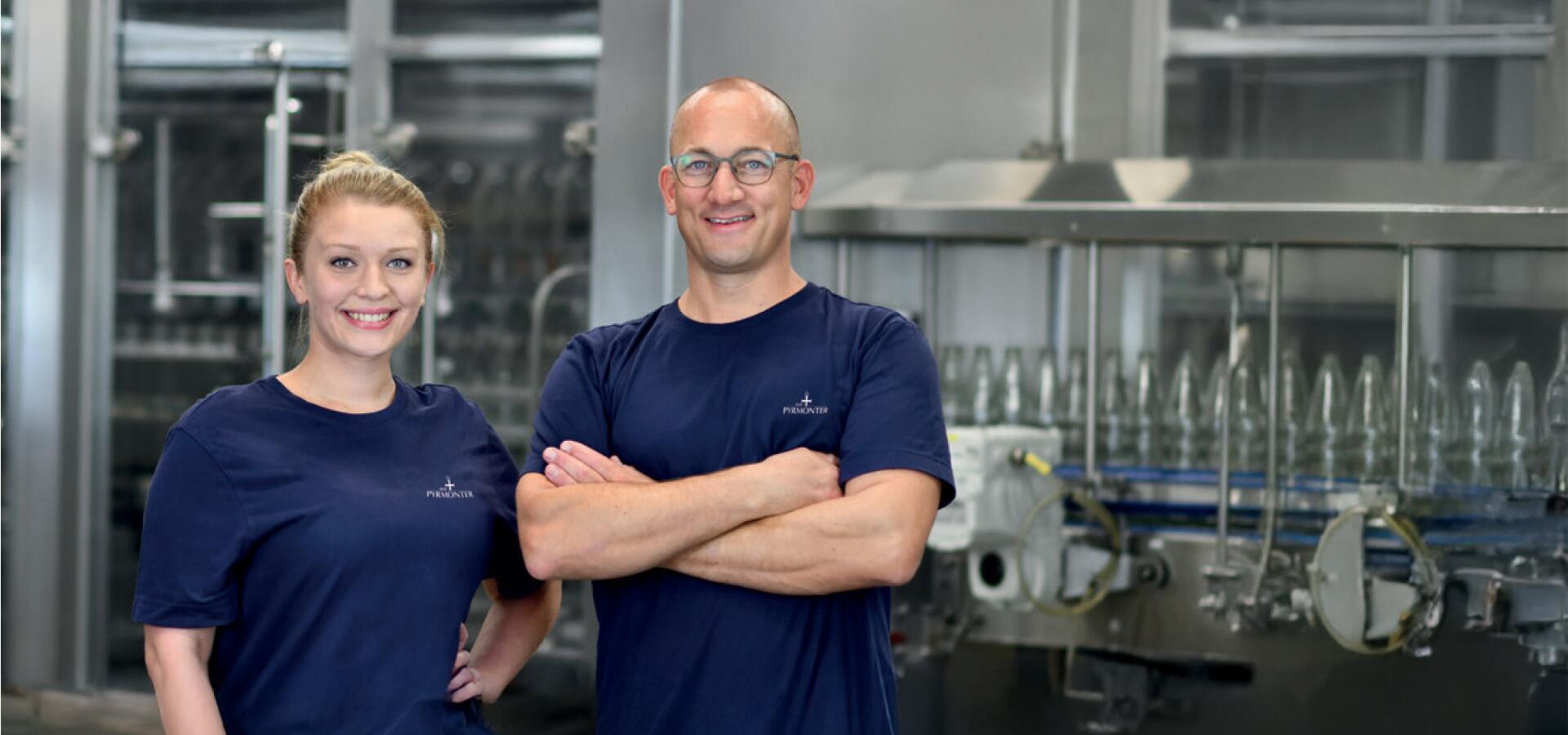 Zwei Mitarbeitende in blauen Bad Pyrmonter T-Shirts stehen lächelnd in der Abfüllanlage.