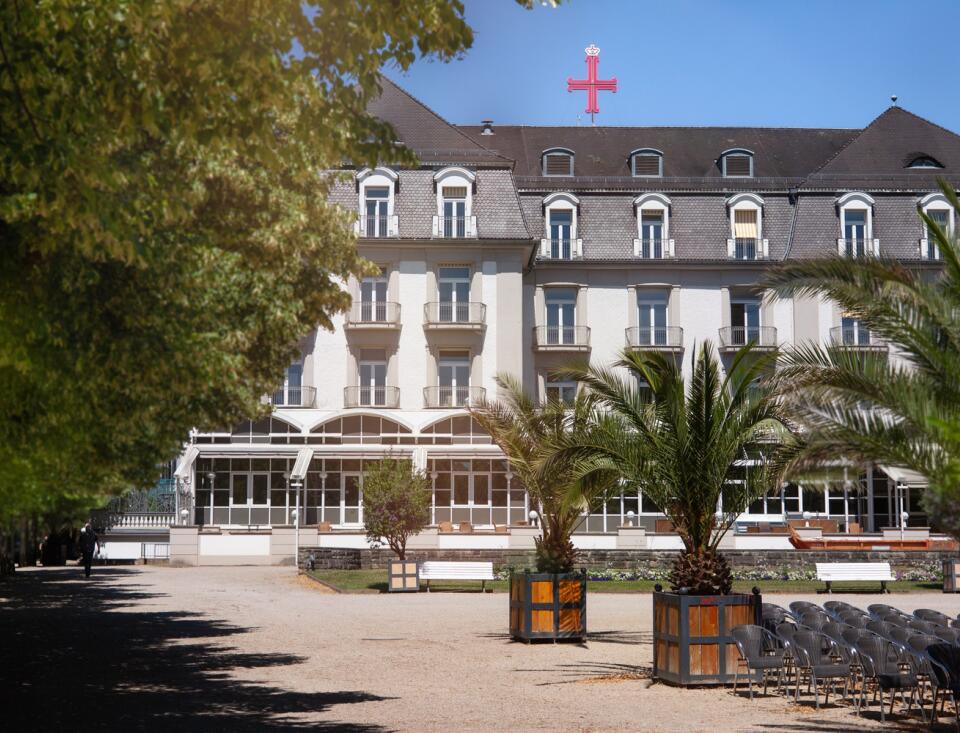 Ein großes, helles Gebäude mit einem grauen Dach, umgeben von grünen Bäumen. Vor dem Gebäude stehen mehrere Palmen in Holzkübeln und leere Stühle. Oben auf dem Dach ist ein rotes Kreuz zu sehen.
