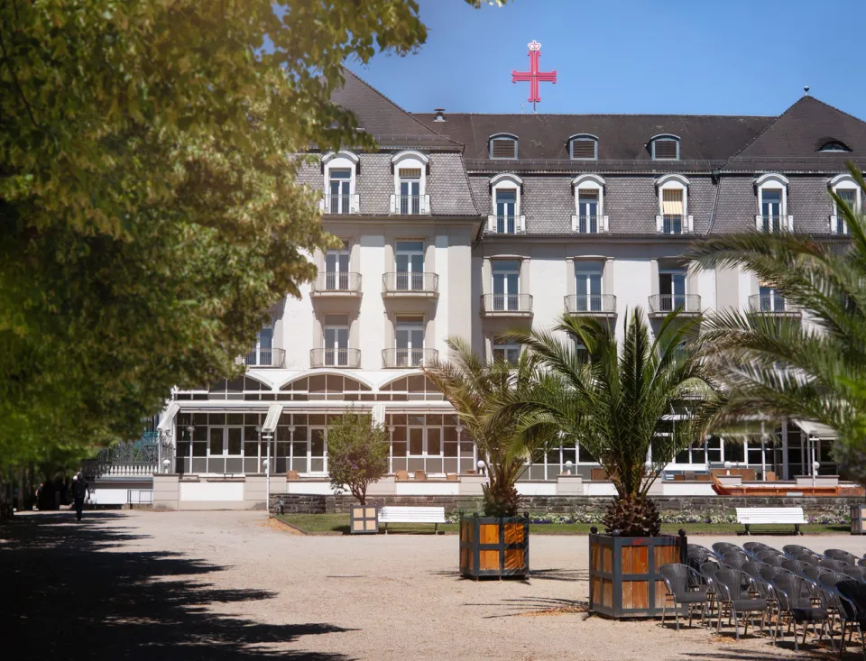 Ein großes, helles Gebäude mit einem grauen Dach, umgeben von grünen Bäumen. Vor dem Gebäude stehen mehrere Palmen in Holzkübeln und leere Stühle. Oben auf dem Dach ist das rote Bad Pyrmont(er) Kreuz zu sehen.