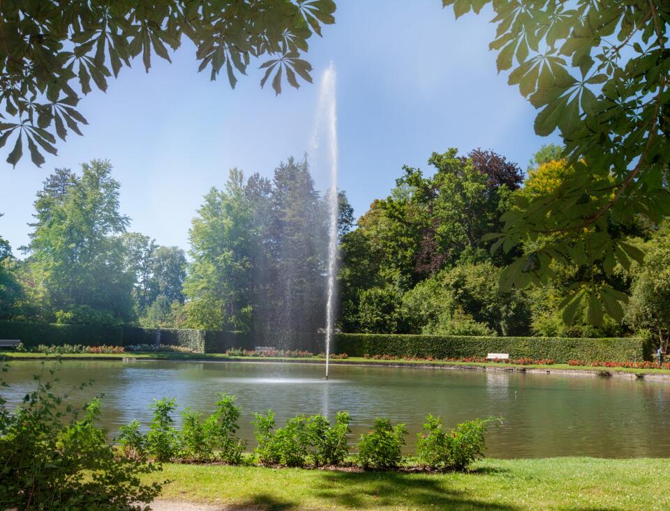 Ein See in einem Park, mit einer Wasserfontäne in der Mitte. Man sieht einen strahlend blauer Himmel und grüne Bäumer drumherum.