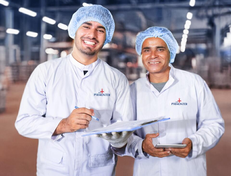 Zwei Mitarbeiter in Hygienekleidung lächeln in die Kamera und überprüfen gemeinsam Unterlagen in einer Produktionshalle von Bad Pyrmont.