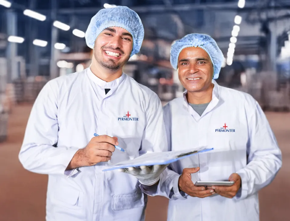 Zwei Mitarbeiter in Hygienekleidung lächeln in die Kamera und überprüfen gemeinsam Unterlagen in einer Produktionshalle von Bad Pyrmont.