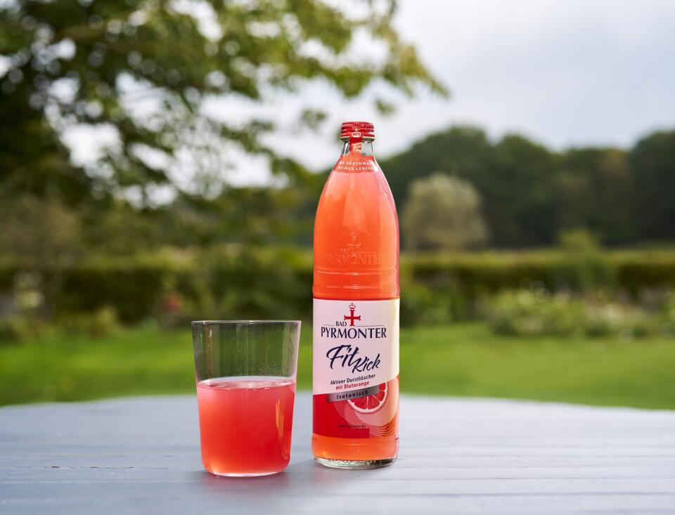 Bad Pyrmonter Fit Kick Blutorange in einer Flasche und in einem Glas stehen auf einem Holztisch vor Naturhintergrund.