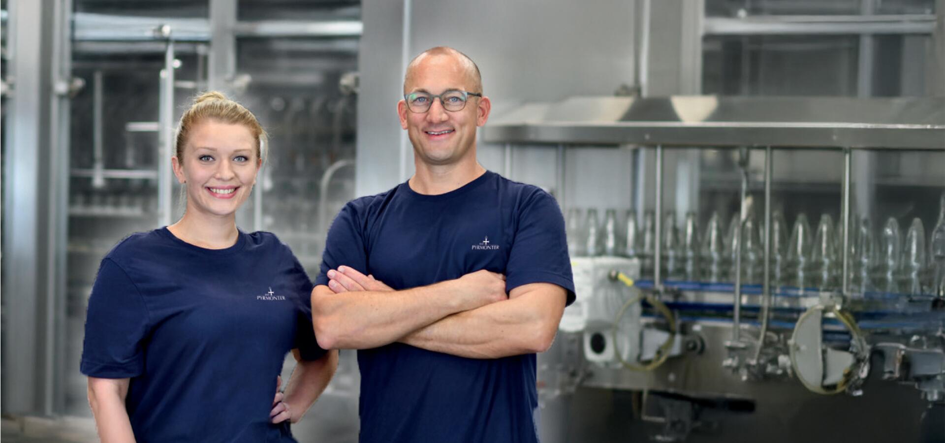 Zwei Mitarbeitende in blauen Bad Pyrmonter T-Shirts stehen lächelnd in der Abfüllanlage.