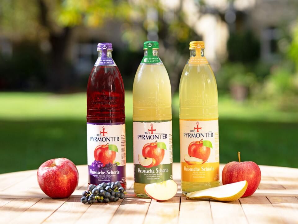 Drei Flaschen Bad Pyrmonter Heimische Schorle in den Sorten Traube, Apfel und Apfel naturtrüb mit frischen Früchten.