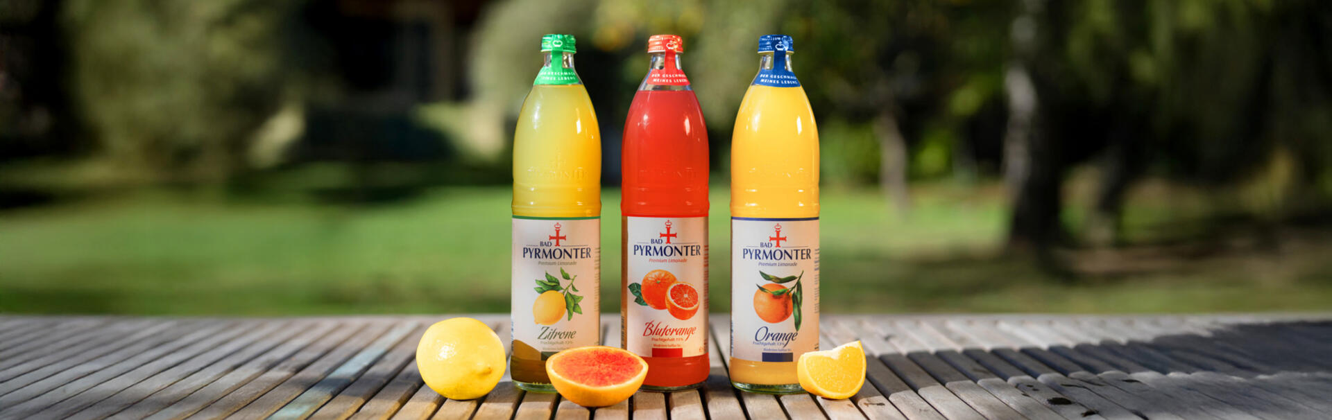 Drei Flaschen Bad Pyrmonter Premium Limonade in den Sorten Zitrone, Blutorange und Orange mit frischen Früchten.