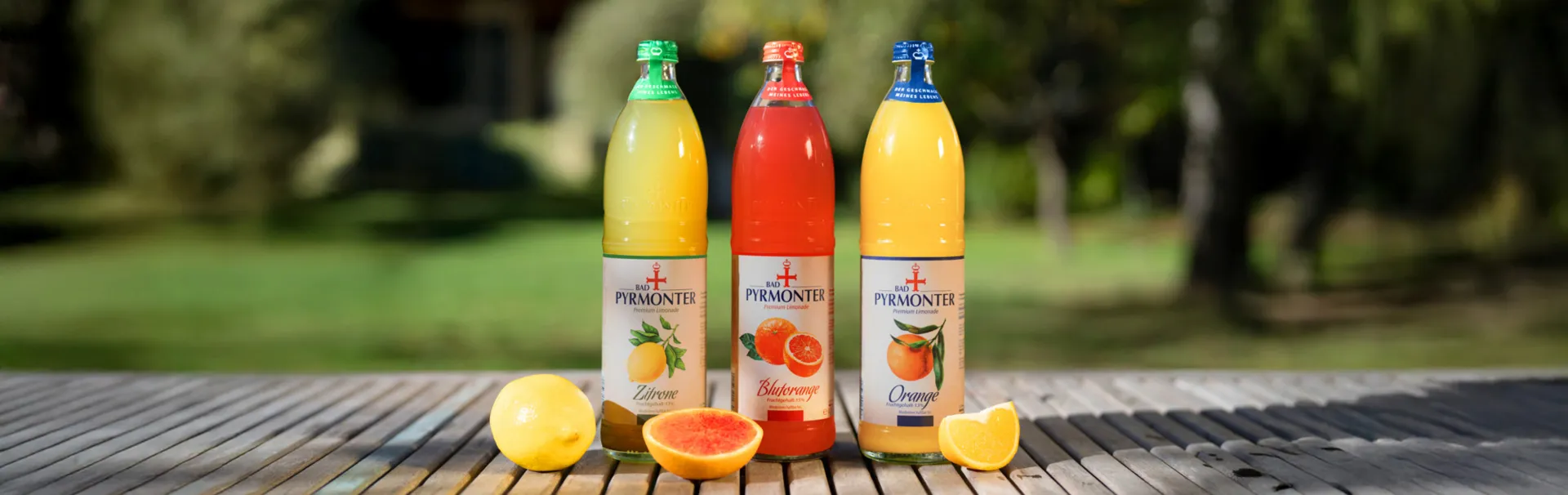 Drei Flaschen Bad Pyrmonter Premium Limonade in den Sorten Zitrone, Blutorange und Orange mit frischen Früchten.