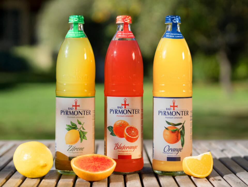 Drei Flaschen Bad Pyrmonter Premium Limonade in den Sorten Zitrone, Blutorange und Orange mit frischen Früchten.