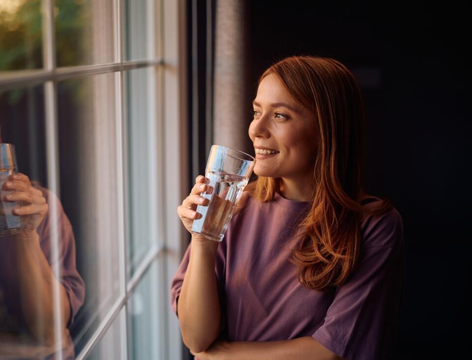 Frau trinkt lächelnd ein Glas Wasser und schaut dabei aus einem Fenster.
