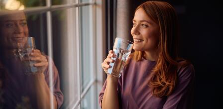 Frau trinkt lächelnd ein Glas Wasser und schaut dabei aus einem Fenster.