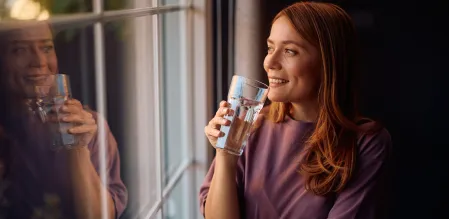 Frau trinkt lächelnd ein Glas Wasser und schaut dabei aus einem Fenster.