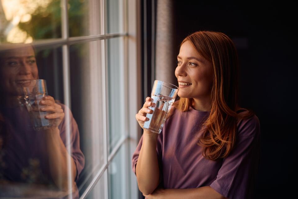 Frau trinkt lächelnd ein Glas Wasser und schaut dabei aus einem Fenster.