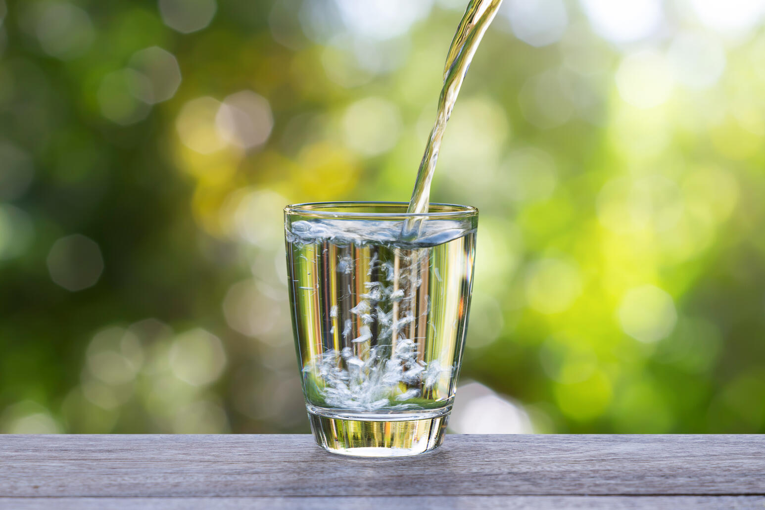 Klares Wasser wird in ein Glas auf einem Holztisch gegossen, mit grünem, sonnendurchflutetem Hintergrund.