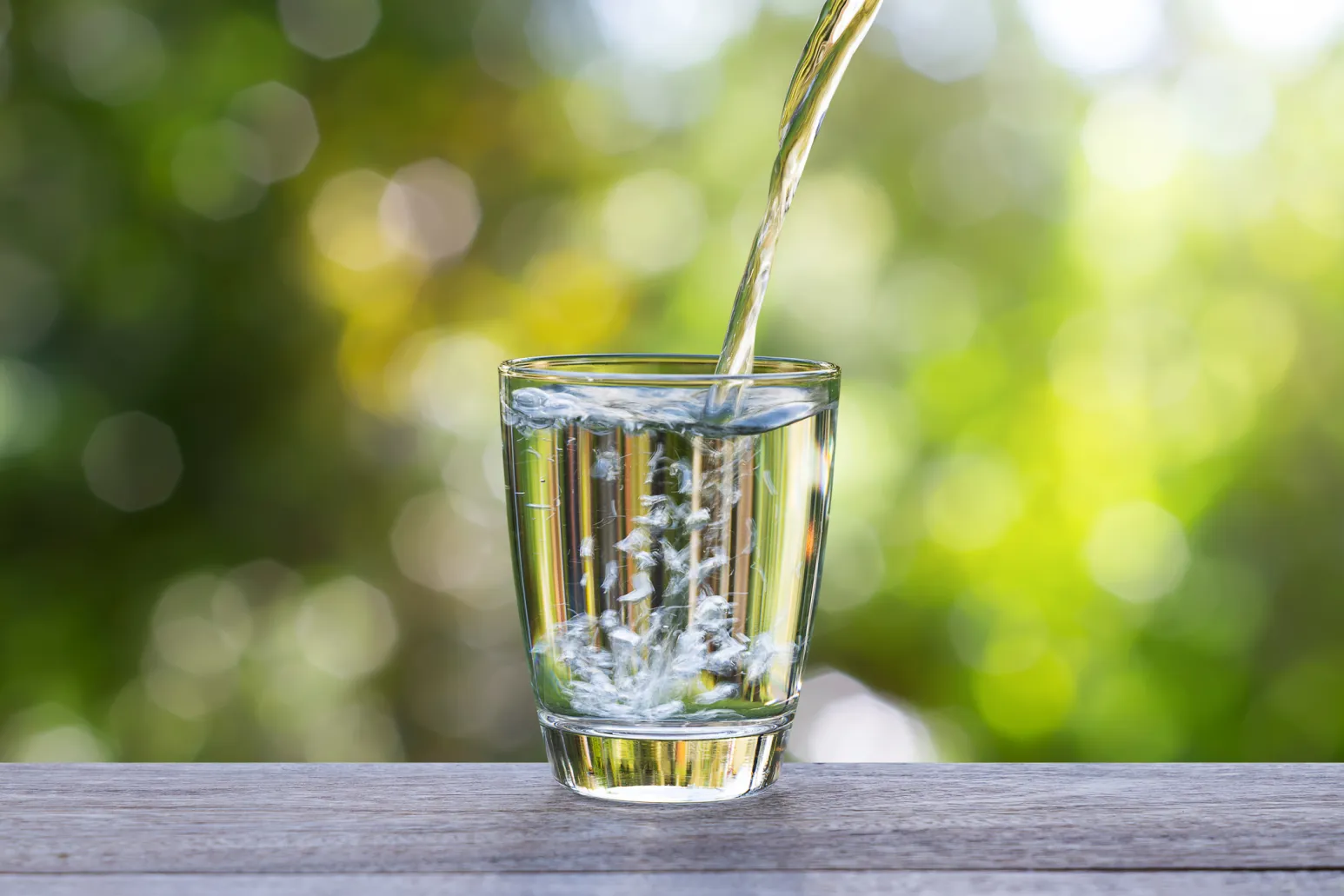 Klares Wasser wird in ein Glas auf einem Holztisch gegossen, mit grünem, sonnendurchflutetem Hintergrund.