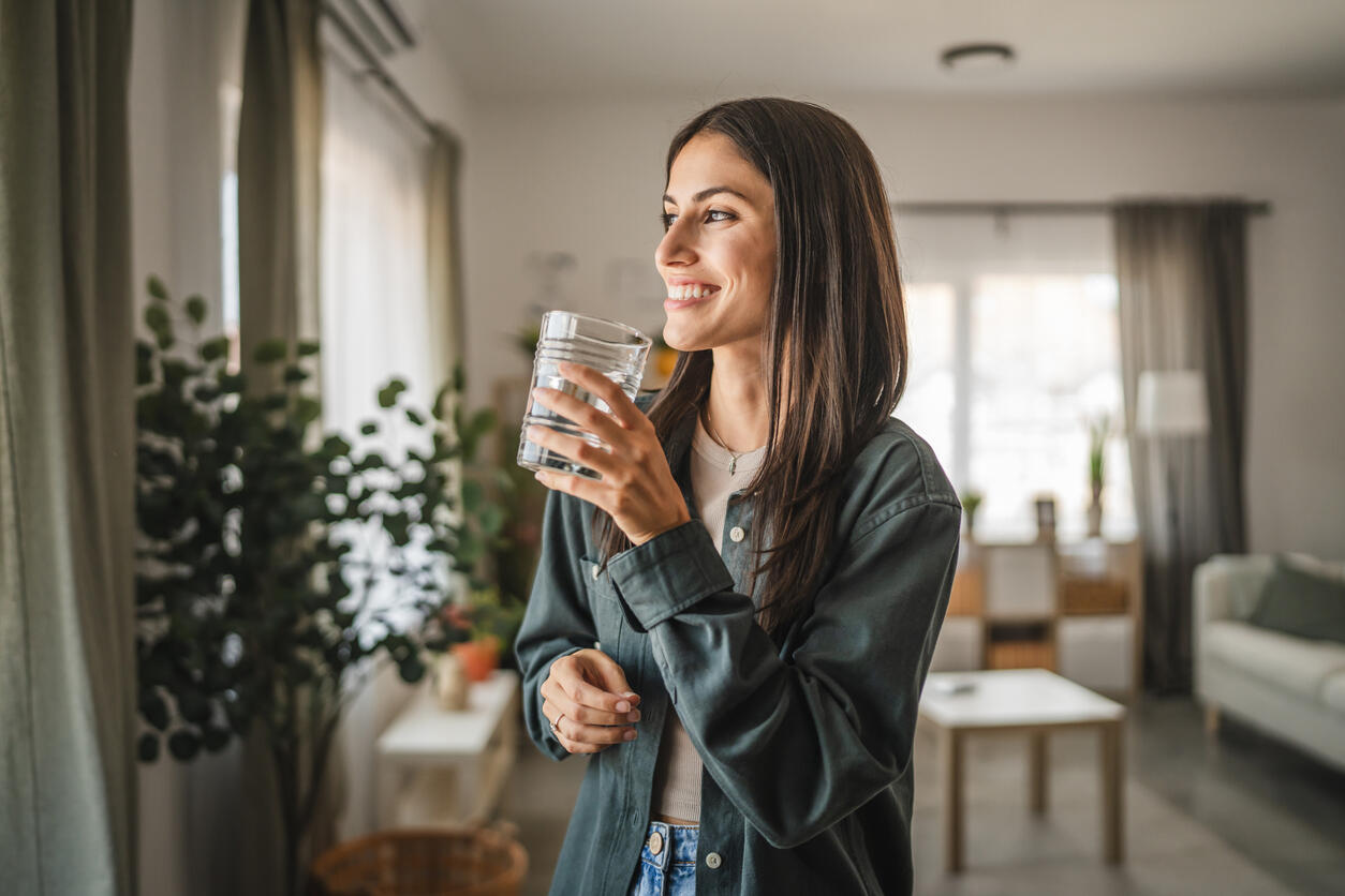Erwachsene Frau steht in einem hellen Wohnraum, hält ein Glas Wasser in der Hand und blickt lächelnd aus dem Fenster. Im Hintergrund sind Pflanzen, ein Tisch und eine Couch zu sehen.