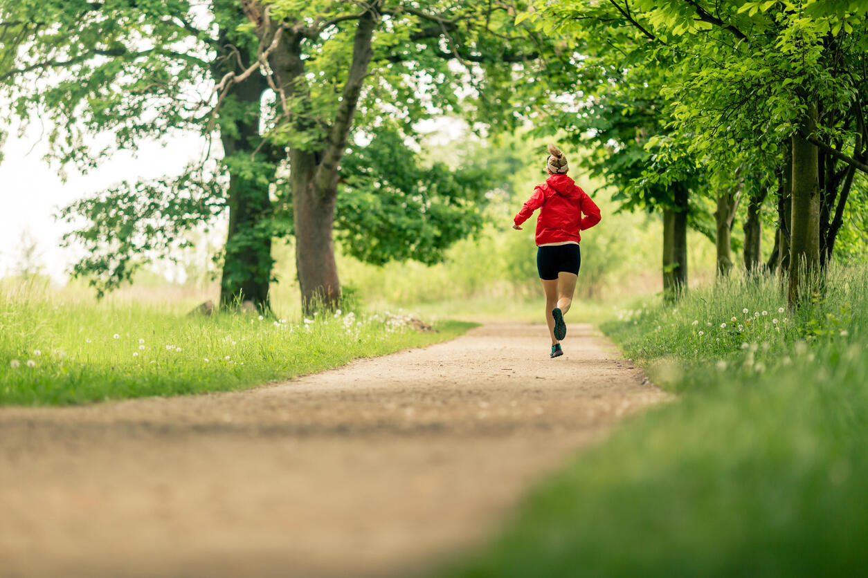 Person joggt von hinten gesehen auf einem naturbelassenen Weg durch eine grüne Park- oder Waldlandschaft. Sie trägt eine rote Jacke und schwarze Sportkleidung, umgeben von Bäumen und Wiesen.