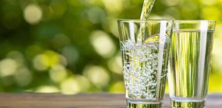 Zwei Wassergläser stehen auf einem Holztisch in grüner Naturumgebung. Ein Glas enthält stilles Wasser, das andere sprudelndes Wasser, in das gerade Wasser eingegossen wird. Im Hintergrund sind unscharf Pflanzen und Grünflächen zu sehen.