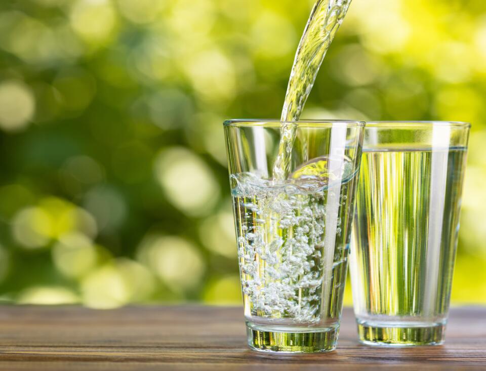 Zwei Wassergläser stehen auf einem Holztisch in grüner Naturumgebung. Ein Glas enthält stilles Wasser, das andere sprudelndes Wasser, in das gerade Wasser eingegossen wird. Im Hintergrund sind unscharf Pflanzen und Grünflächen zu sehen.