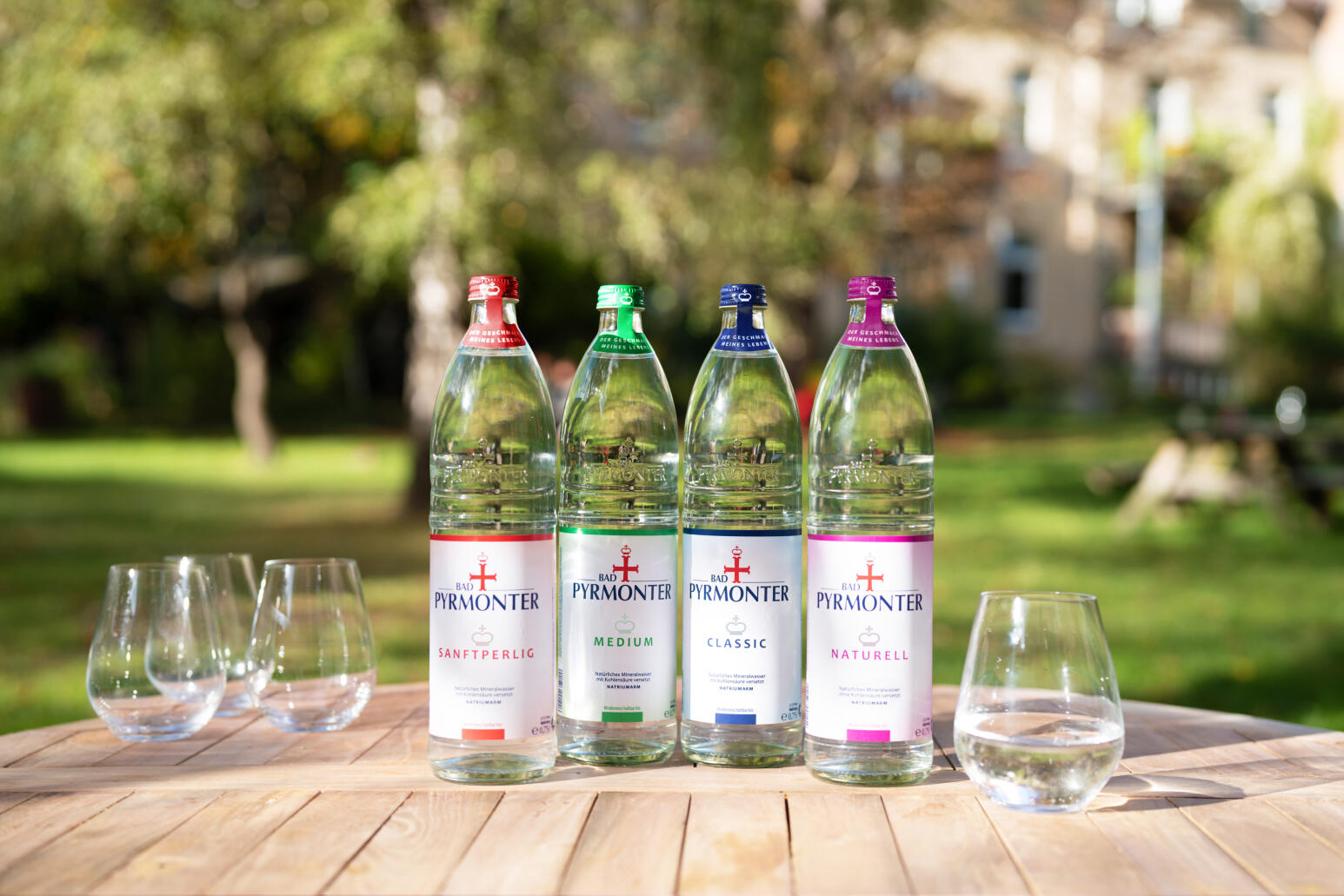 Bad Pyrmonter Mineralwasser Range steht in Glasflaschen auf einem Gartentisch aus Holz. Links daneben stehen drei Gläser, rechts eins. Im Hintergrund sieht man einen Garten mit grünen Bäumen.