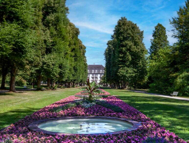 Aufnahme aus dem Kurpark in Bad Pyrmont. Im Vordergrund sieht man lilane Blumen und ein Wasserbecken mit einer Fontäne. Im Hintergrund sind viele grüne Bäume, sowie das Schloss Pyrmont mit dem roten Pyrmont Kreuz auf dem Dach.