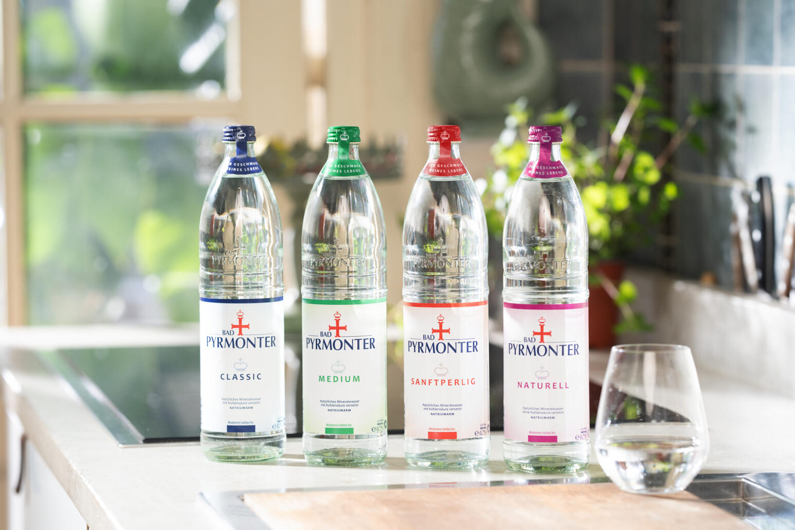 Vier Flaschen Bad Pyrmonter Mineralwasser in den Sorten Classic, Medium, Sanftperlig und Naturell stehen nebeneinander auf einer hellen Küchenarbeitsfläche; rechts daneben steht ein gefülltes Trinkglas.