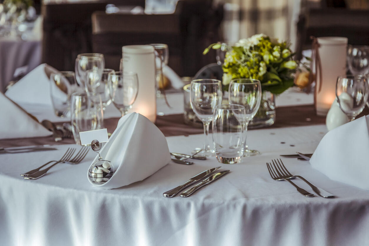Festlich gedeckter runder Tisch mit weißer Tischdecke, gefalteten weißen Servietten, mehreren Wasser- und Weingläsern, Besteck sowie einem niedrigen Blumenarrangement in der Tischmitte.