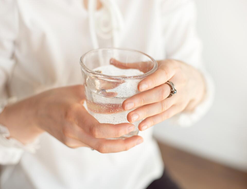 Nahaufnahme einer Person in heller Kleidung, die ein Glas mit kohlensäurehaltigem Wasser mit beiden Händen hält; im Wasser sind feine aufsteigende Bläschen zu erkennen.