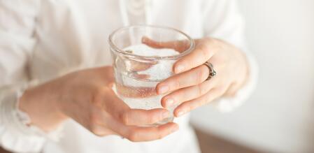 Nahaufnahme einer Person in heller Kleidung, die ein Glas mit kohlensäurehaltigem Wasser mit beiden Händen hält; im Wasser sind feine aufsteigende Bläschen zu erkennen.