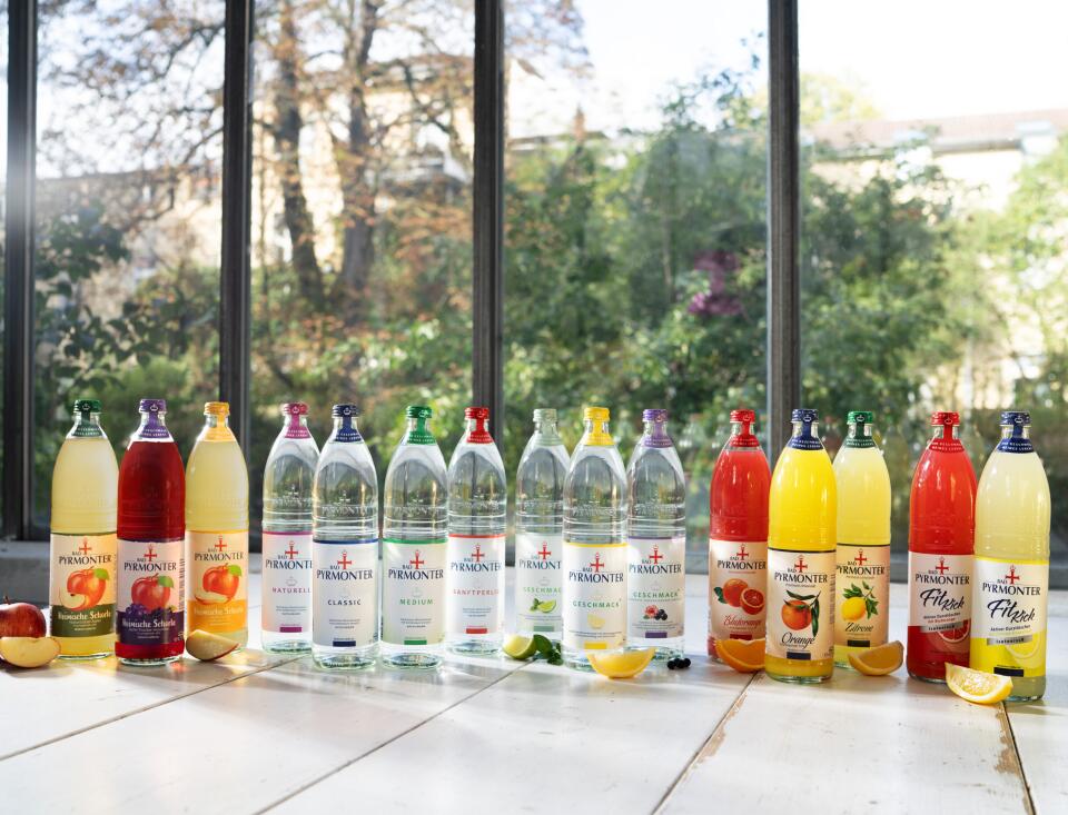 Mehrere Flaschen Bad Pyrmonter Getränke und Mineralwasser stehen in einer Reihe auf einem hellen Holztisch vor einer großen Fensterfront; gezeigt werden verschiedene Sorten, darunter Schorlen, Mineralwasser und Erfrischungsgetränke, ergänzt durch aufgeschnittenes Obst wie Apfel, Zitrone und Orange.
