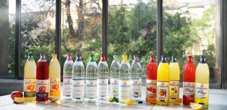 Mehrere Flaschen Bad Pyrmonter Getränke und Mineralwasser stehen in einer Reihe auf einem hellen Holztisch vor einer großen Fensterfront; gezeigt werden verschiedene Sorten, darunter Schorlen, Mineralwasser und Erfrischungsgetränke, ergänzt durch aufgeschnittenes Obst wie Apfel, Zitrone und Orange.