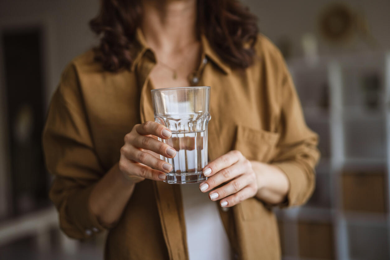 Person in brauner Bluse hält in einem Innenraum ein gefülltes Wasserglas mit beiden Händen auf Brusthöhe.