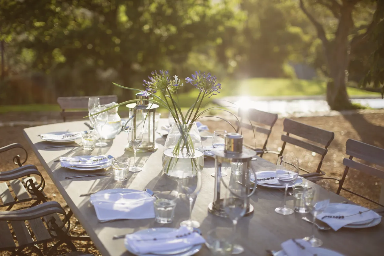 Langer, festlich gedeckter Tisch im Freien mit Tellern, Gläsern, weißen Servietten und Laternen; in der Mitte steht eine Vase mit violetten Blumen, im Hintergrund ist ein sonniger Garten zu sehen.