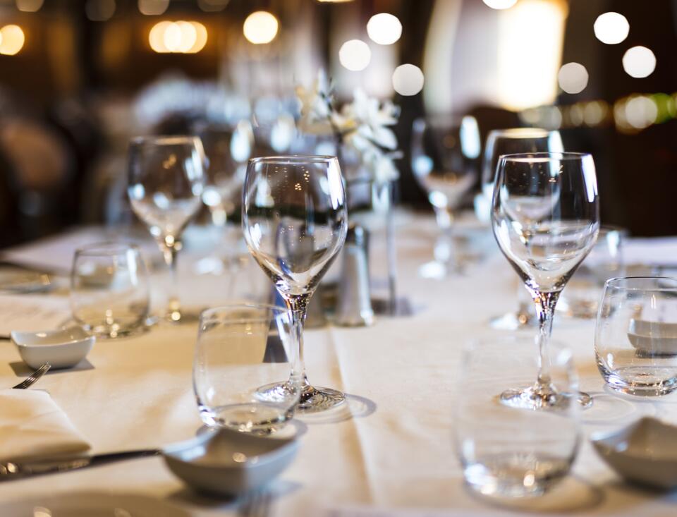 Gedeckter Restauranttisch mit mehreren Wasser- und Weingläsern auf weißer Tischdecke; im Hintergrund warme, unscharfe Lichter und eine elegante Atmosphäre.