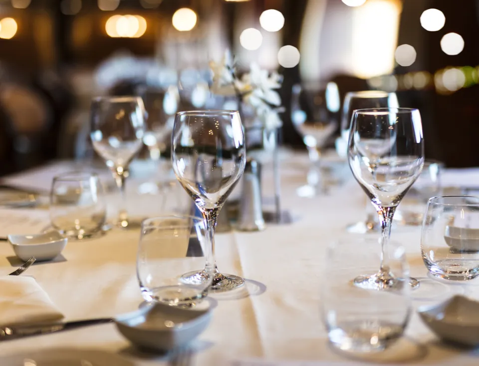 Gedeckter Restauranttisch mit mehreren Wasser- und Weingläsern auf weißer Tischdecke; im Hintergrund warme, unscharfe Lichter und eine elegante Atmosphäre.