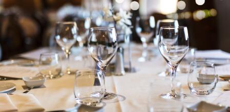 Gedeckter Restauranttisch mit mehreren Wasser- und Weingläsern auf weißer Tischdecke; im Hintergrund warme, unscharfe Lichter und eine elegante Atmosphäre.