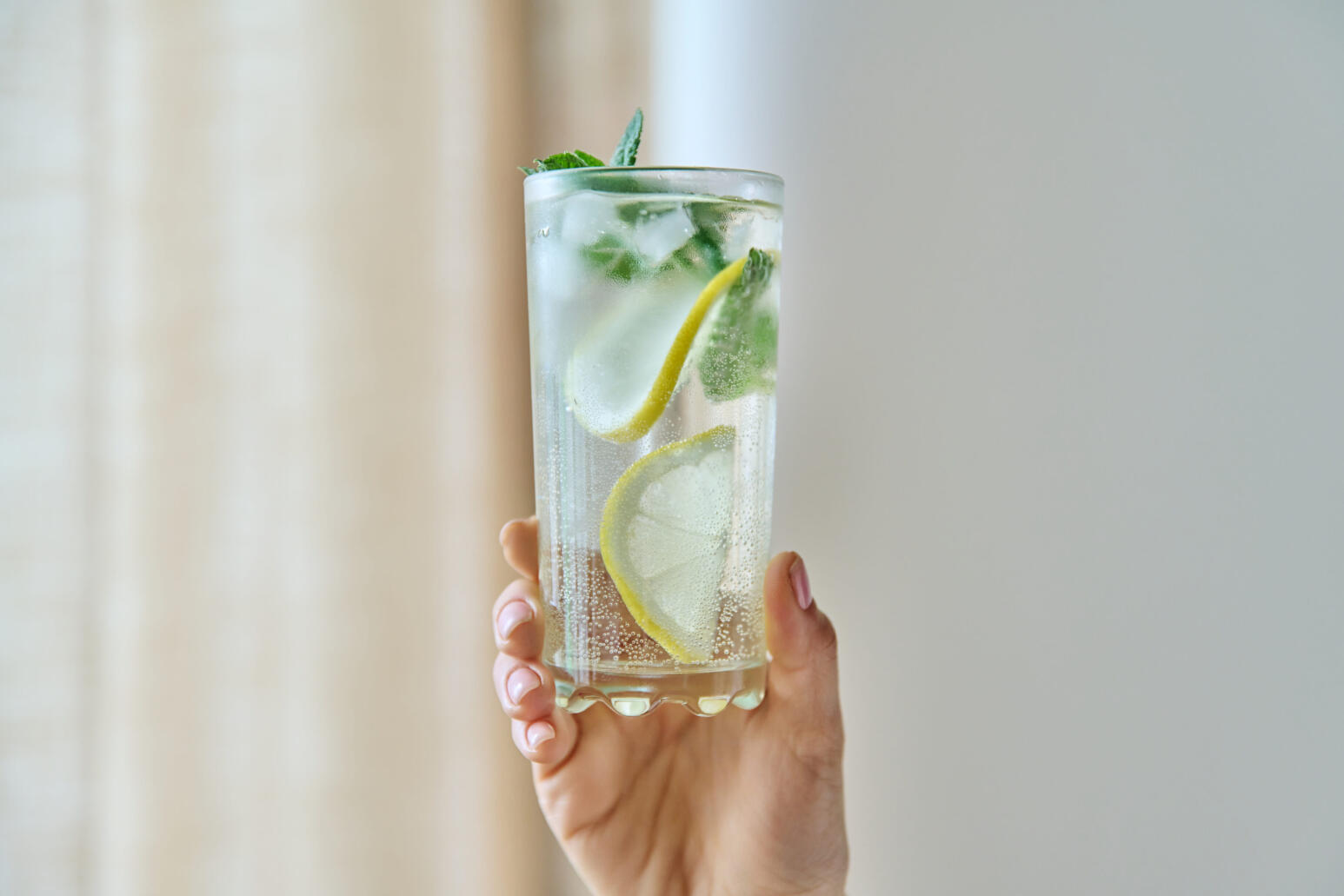Hand hält ein hohes Glas mit sprudelndem Wasser, Eiswürfeln, Zitronenscheiben und frischer Minze vor hellem, unscharfem Hintergrund.