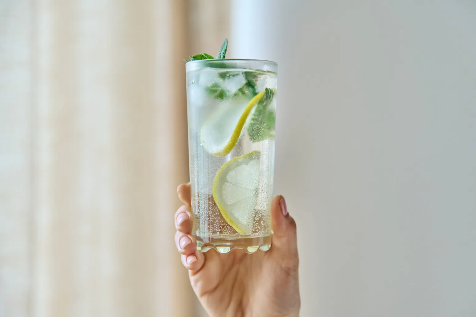 Hand hält ein hohes Glas mit sprudelndem Wasser, Eiswürfeln, Zitronenscheiben und frischer Minze vor hellem, unscharfem Hintergrund.