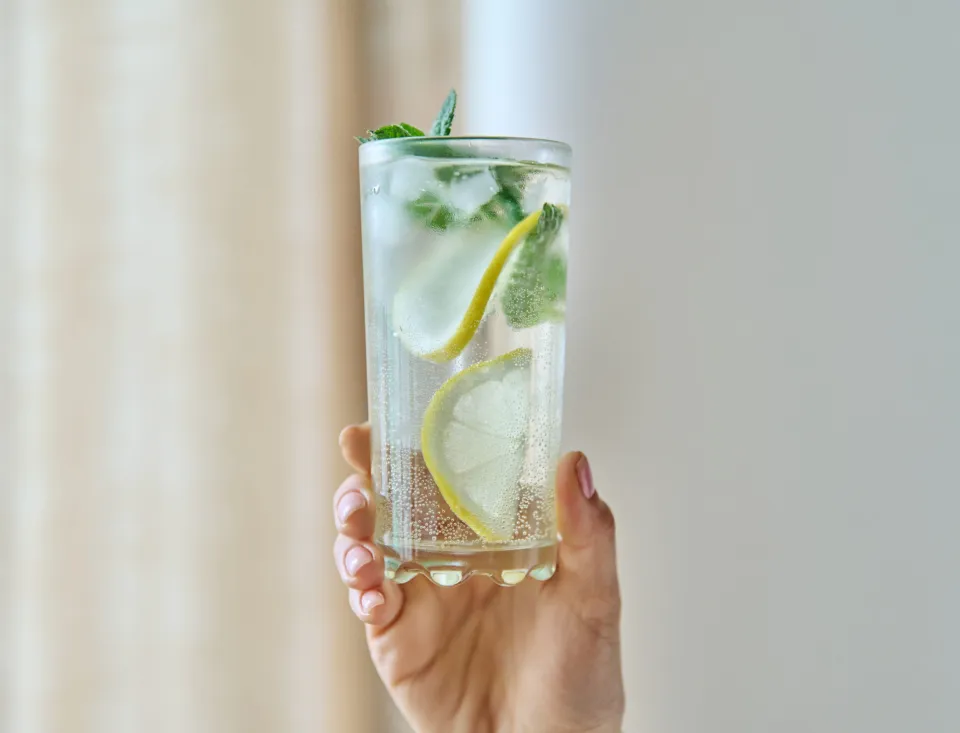 Hand hält ein hohes Glas mit sprudelndem Wasser, Eiswürfeln, Zitronenscheiben und frischer Minze vor hellem, unscharfem Hintergrund.