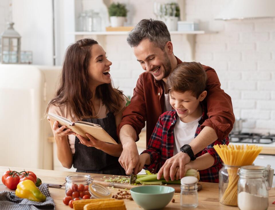 Eine Familie bereitet gemeinsam in einer hellen Küche eine Mahlzeit zu. Die Mutter hält ein Kochbuch und lacht, während Vater und Sohn zusammen Gemüse auf einem Schneidebrett schneiden. Auf der Arbeitsfläche liegen frische Zutaten wie Tomaten, Paprika, Maiskolben, Sellerie und Spaghetti in Gläsern.
