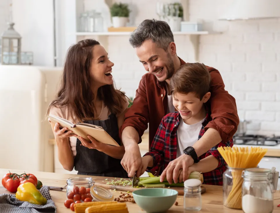 Eine Familie bereitet gemeinsam in einer hellen Küche eine Mahlzeit zu. Die Mutter hält ein Kochbuch und lacht, während Vater und Sohn zusammen Gemüse auf einem Schneidebrett schneiden. Auf der Arbeitsfläche liegen frische Zutaten wie Tomaten, Paprika, Maiskolben, Sellerie und Spaghetti in Gläsern.