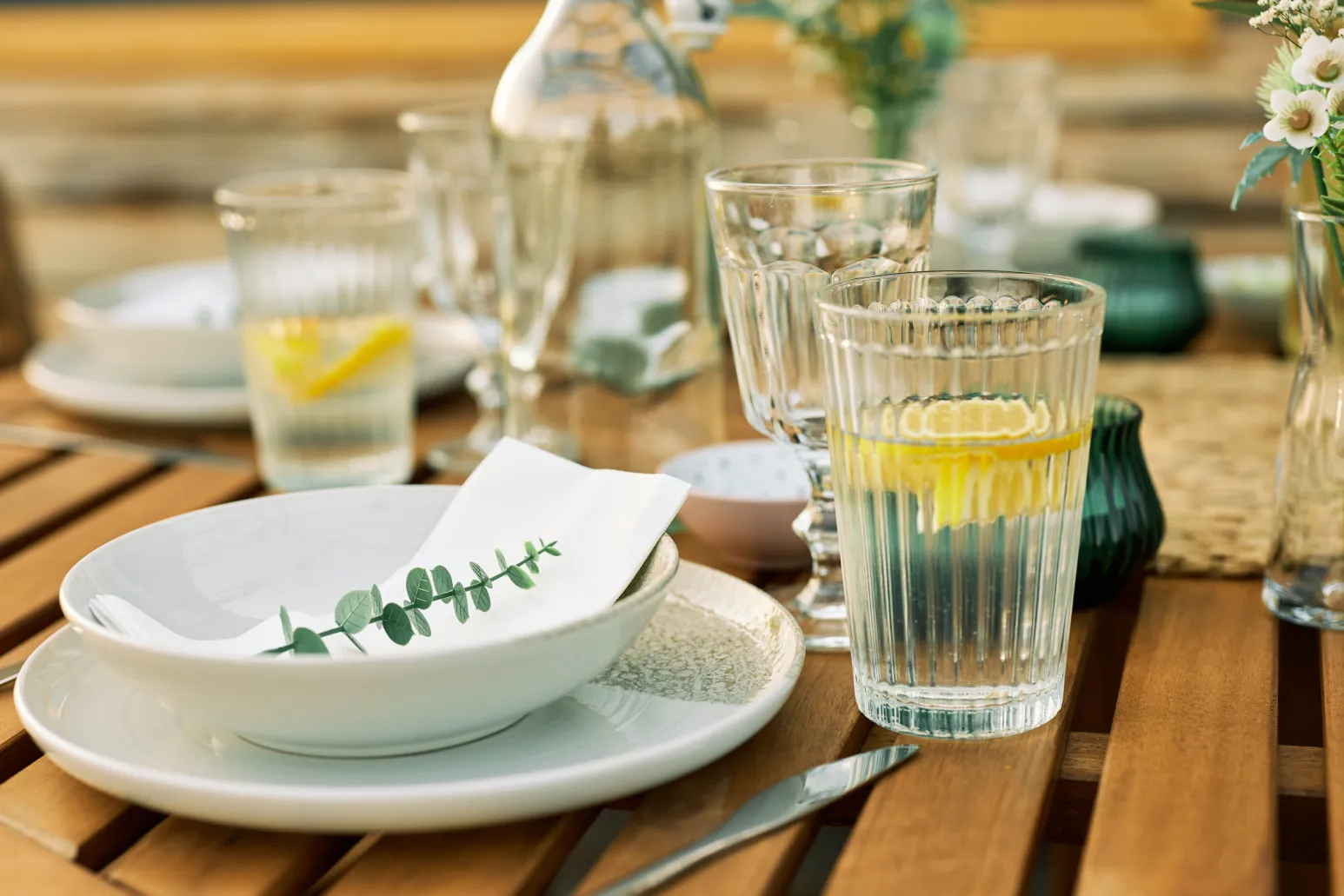 Auf einem gedeckten Holztisch stehen mehrere Gläser mit Wasser, teils mit Zitronenscheiben, neben Tellern, Besteck und kleinen Blumen.
