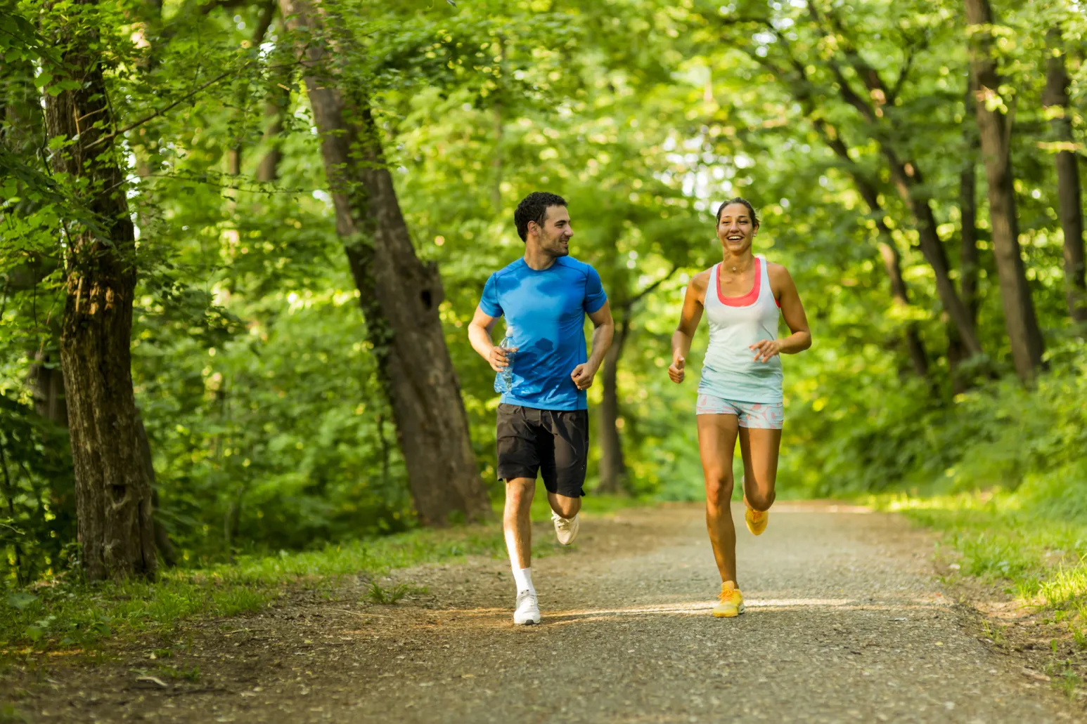 Eine weibliche und eine männliche Person joggen gemeinsam auf einem breiten Waldweg durch einen dicht begrünten, sonnigen Laubwald; eine Person trägt eine Wasserflasche in der Hand.
