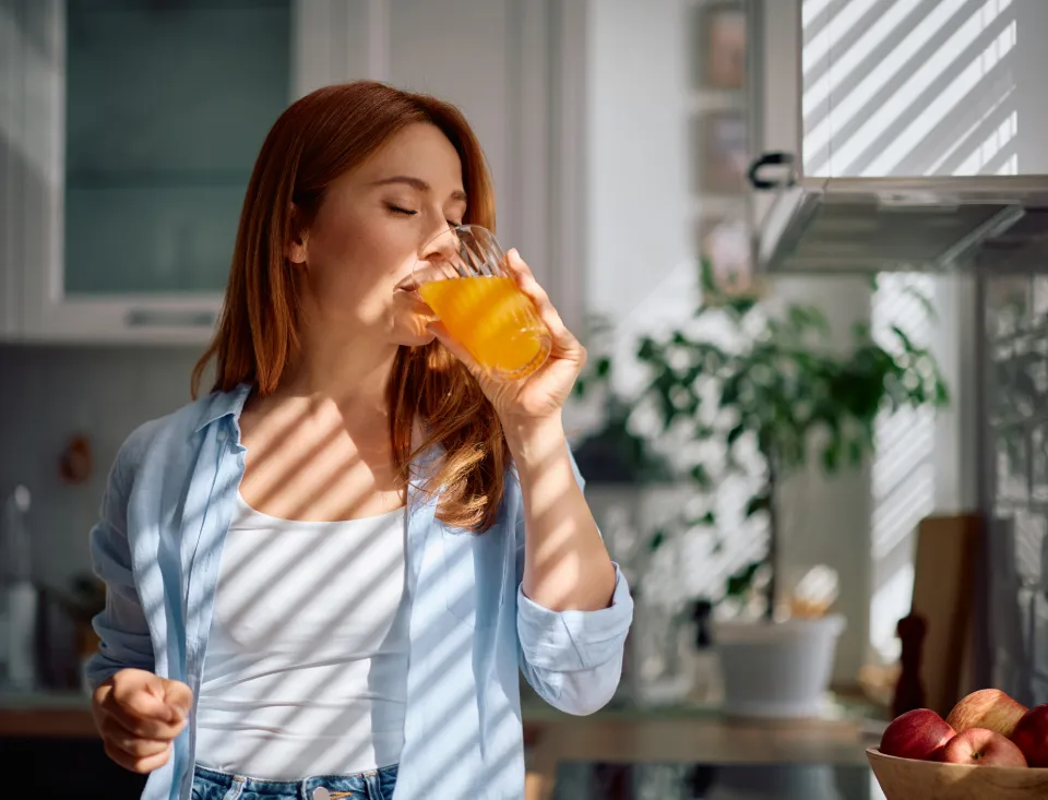 Frau trinkt in einer hellen Küche ein orangefarbenes Getränk aus einem Glas; auf der Arbeitsfläche steht eine Schale mit Äpfeln.