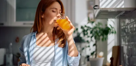 Frau trinkt in einer hellen Küche ein orangefarbenes Getränk aus einem Glas; auf der Arbeitsfläche steht eine Schale mit Äpfeln.