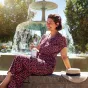 Eine lächelnde Frau sitzt auf einem Brunnenrand. Sie trägt einen rot gemusterten Jumpsuit und hält eine kleine Flasche in der Hand. Ein Strohhut liegt neben ihr.