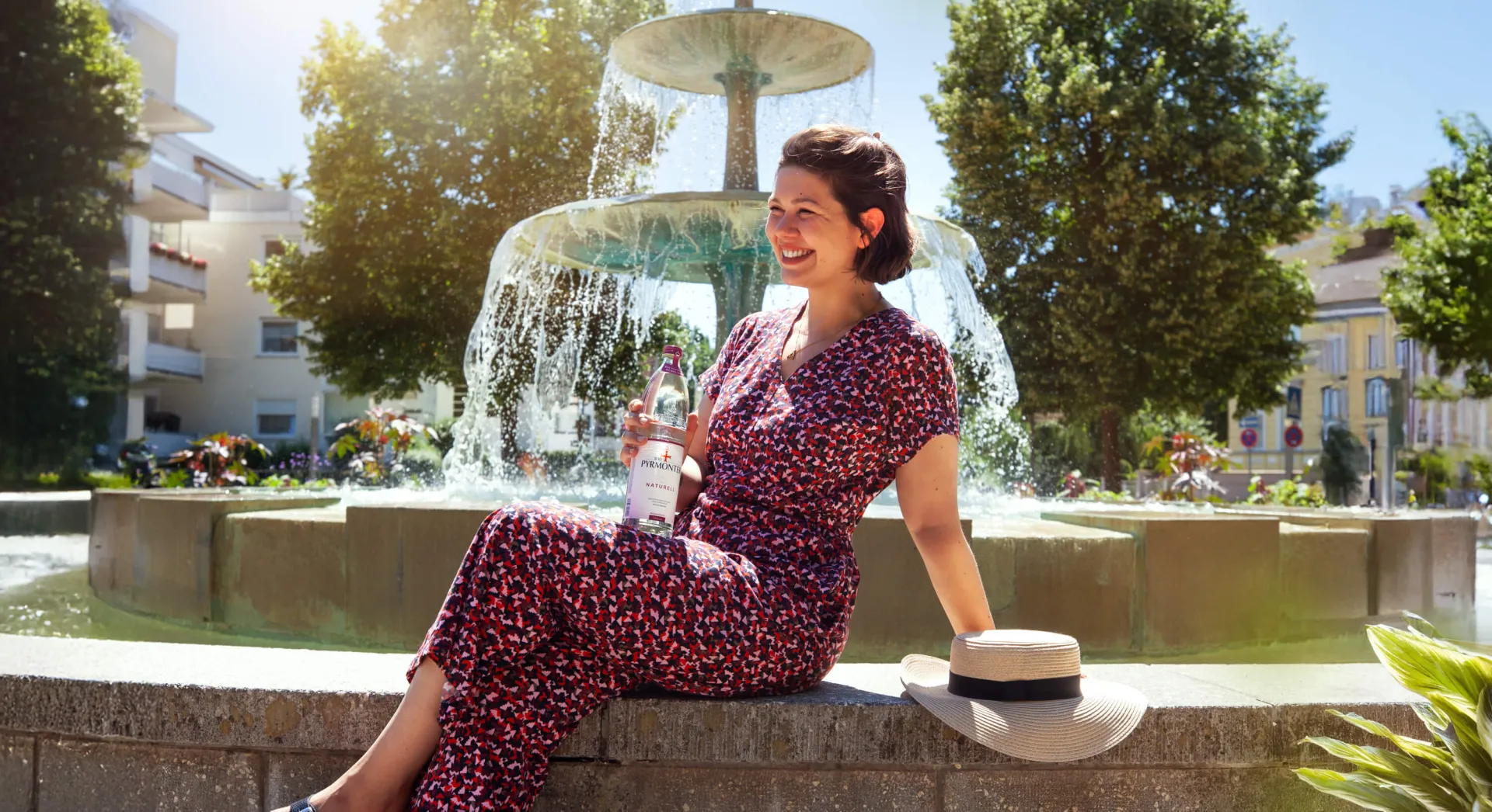 Eine lächelnde Frau sitzt auf einem Brunnenrand. Sie trägt einen rot gemusterten Jumpsuit und hält eine kleine Flasche in der Hand. Ein Strohhut liegt neben ihr.